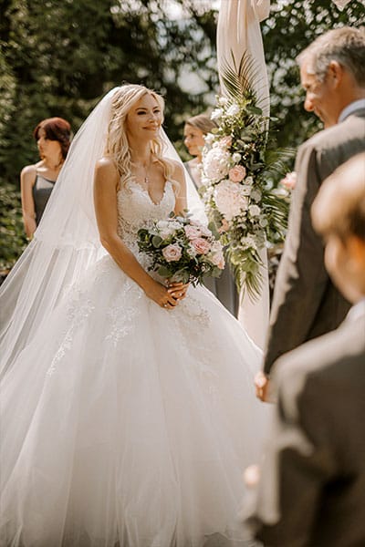 Stunning Bride in a white princess gown with trail and veil stands with her bridal bouquet and looks at her future husband at her wedding ceremony at vila Bled.