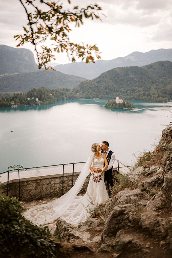 Bride in white gown and groom are hugging at Bled Castle terrace and behind them is lake Bled island with a Church.
