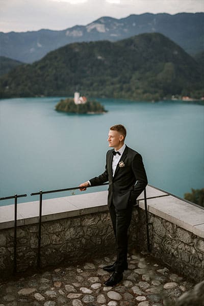 Groom in tailored black suit is looking a Bled town from Lake Bled Casle terrace.