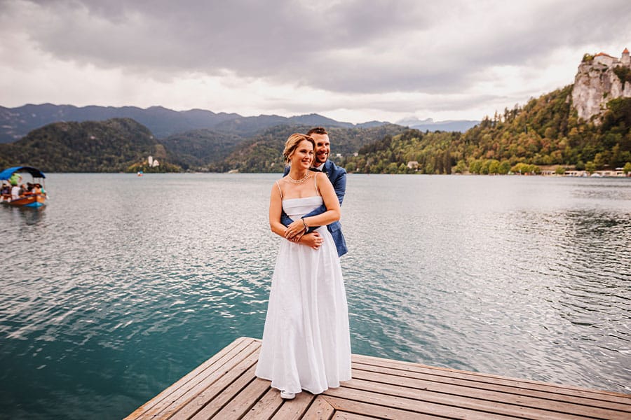 Bride and groom are hugging at Lake Bled pier. Behind them are Lake Bled Island, pletna boat and Bled Castle.