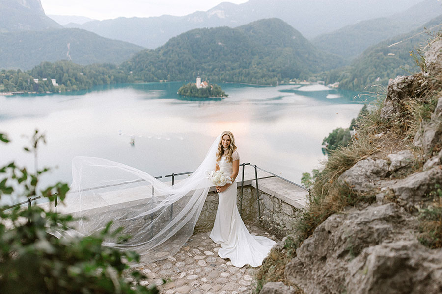 Elegant bride in a flowing gown posing at Bled Castle with a panoramic view of Lake Bled and Bled Island behind her.