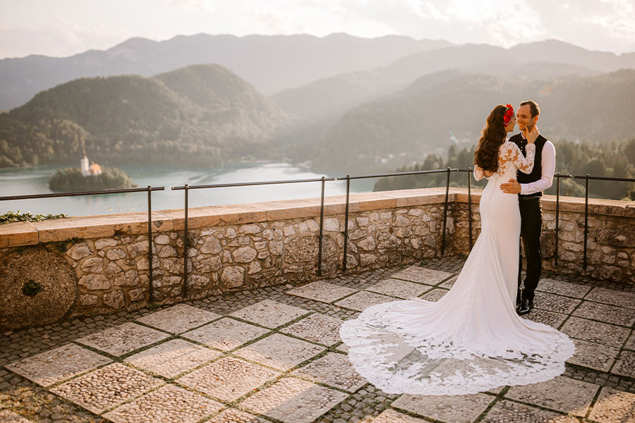 A bride holds her hand on a groom's face with a beautiful view of neighbury mountains, and a Bled lake with a famous island with Bled Church in the warm summer day.