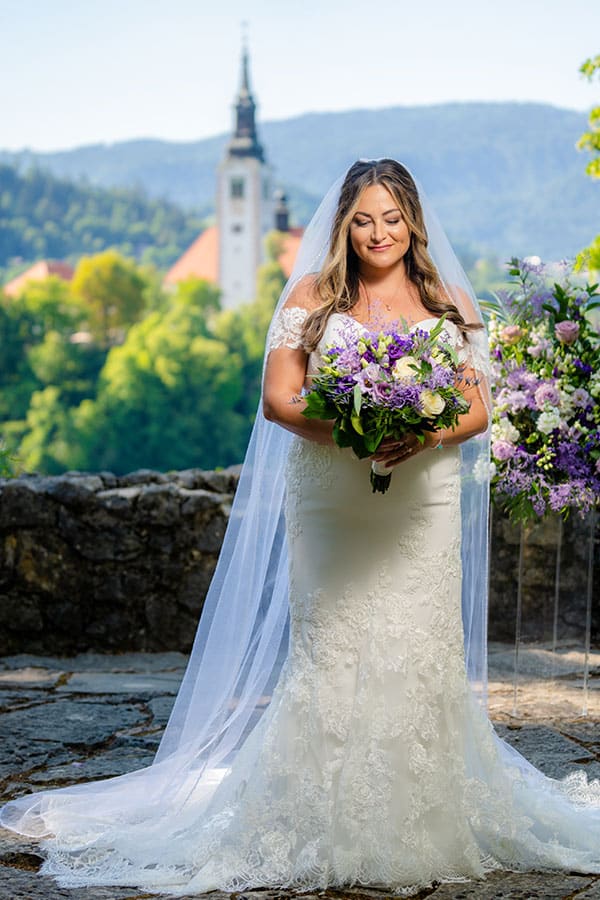 A stunning bride in creamy white lace wedding gown with a long veil and trail looks at her purple bridal bouquet.