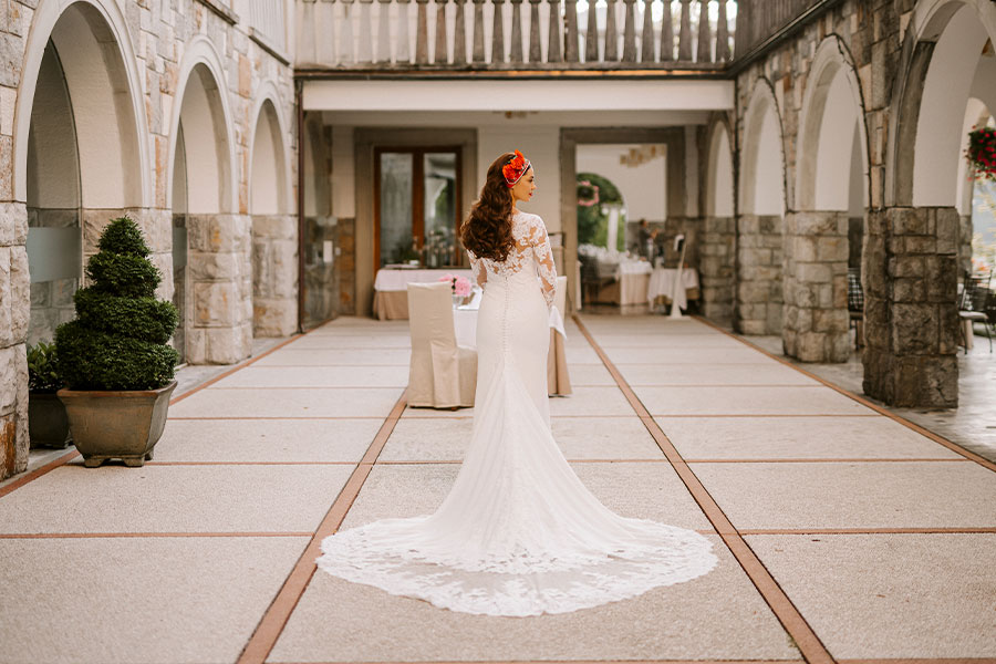 A back view of an amazing bride in a white wedding dress with a long train, and red flowers in her hair stands in an empty villa bled terrace.
