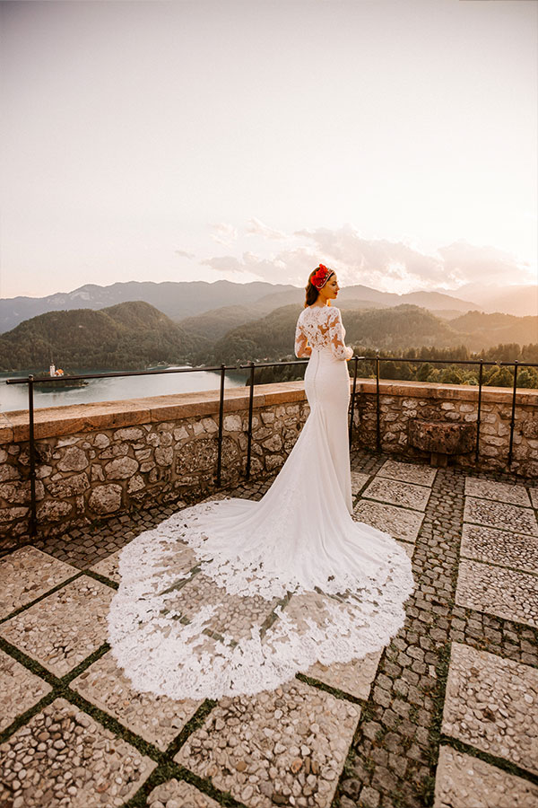 An amazing bride in a wedding dress stands on a beautiful bled castle terrace overlooking mountains, lake Bled island, and lake Bled.