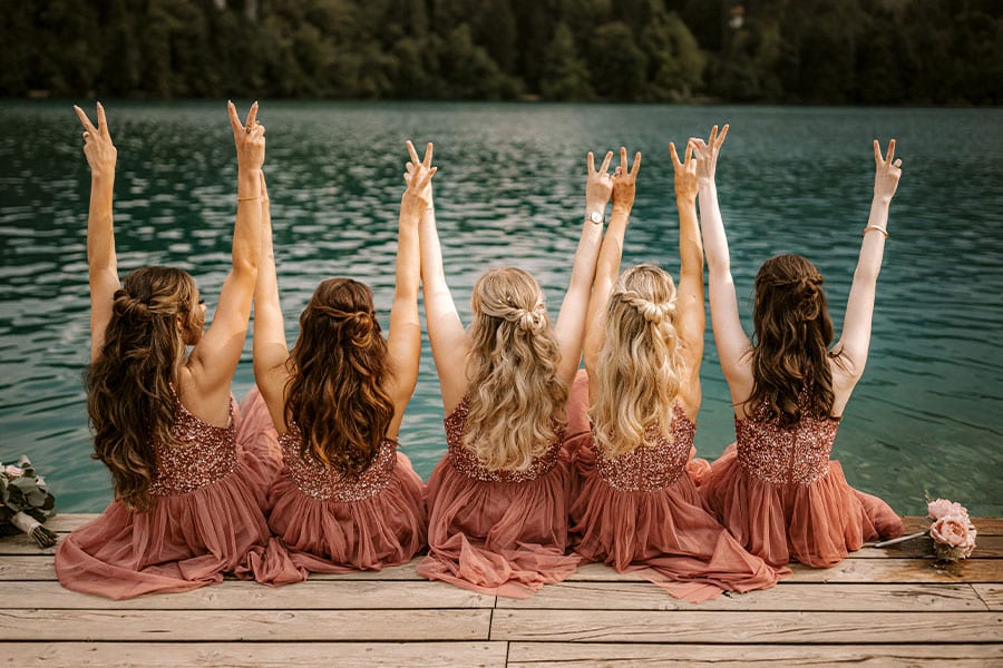 The bridesmaids in their dusty pink dresses sit on the lake Bled pier and hold their hands in the air.