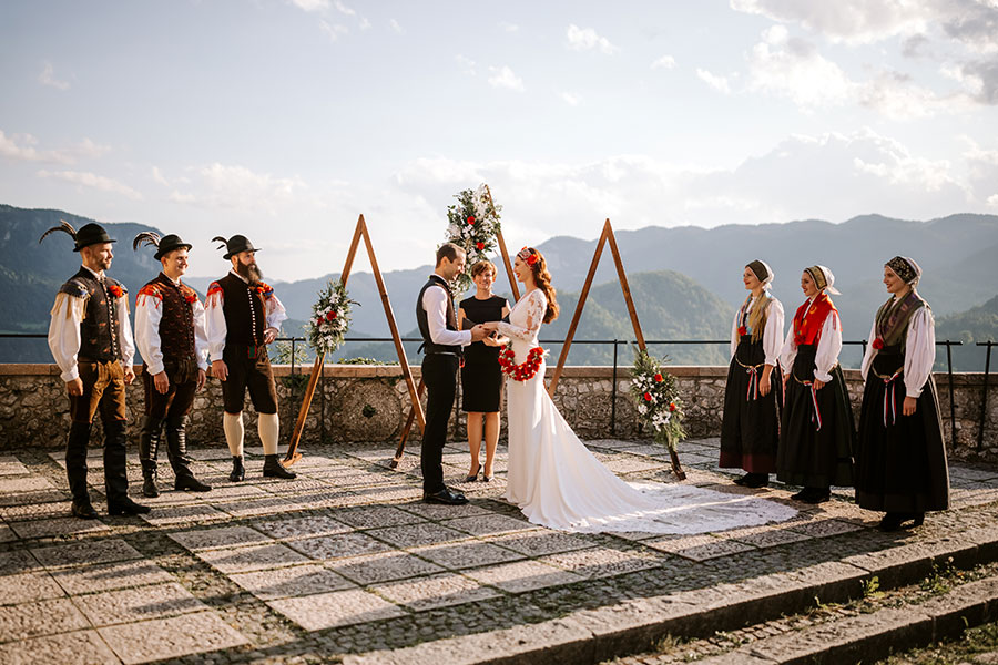 Groom and bride are holding hands during their lake Bled Castle wedding ceremony in front of a wedding officiant and next to folk dancers at their wedding party.