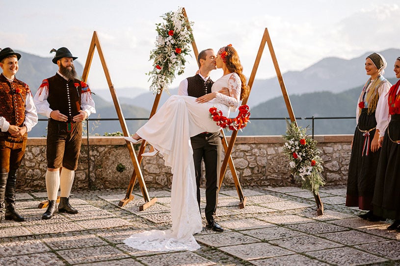 Traditional Slovenian dance group is a wedding party at the wedding of a couple from America at Bled Castle. Groom holds his beautiful bride in a white long dress and red flower crown in his hands and kisses.