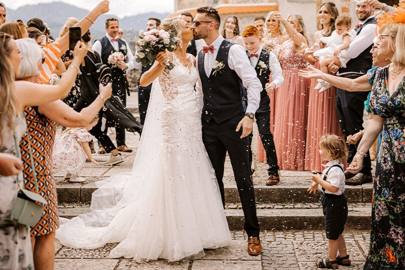 Newlywed kisses during wedding guests throw flower petals at their wedding ceremony in Lake Bled Castle.