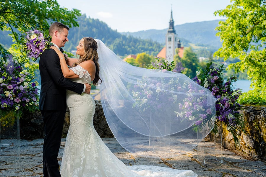 Bride in white long wedding dress with her violet bouquet and groom in a black suit hold each other at ceremony setting at vila Bled ceremony spot and behind them are flower centerpieces on glass stands.