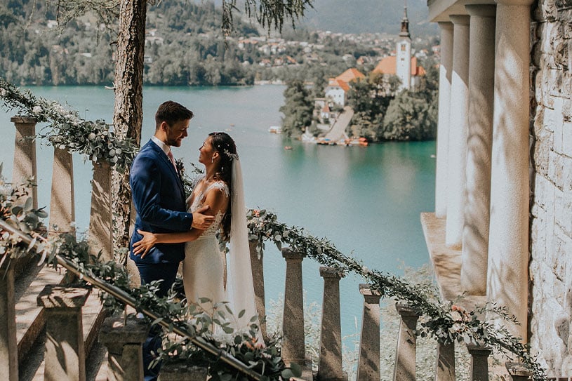 Groom in blue suit and a bride in Pronovias lace dress with a train and veil are hugging on the Pavilion Belvedere stairs next to villa Bled and behind them is Lake Bled Island with a church.
