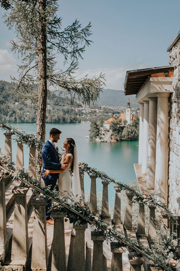 Couple stands on decorated steps with white and green flowers at vila Bled.