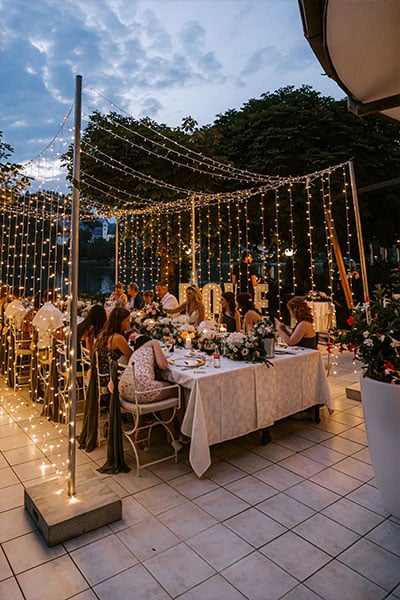 Night photo of wedding guests during dinner at long dinner table decorated with white cloth, pastel color flower garland, candles, silver plates, wooden name tags, wedding menus and personal favours in pink at Grand hotel Toplice, Lake Bled undeer fairy lights tunnel.
