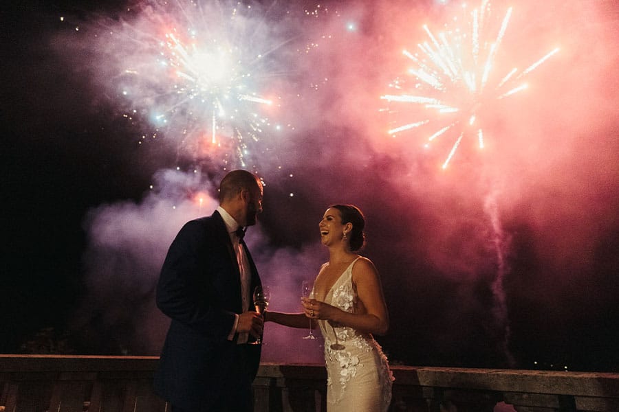 Bride and groom are smiling and holding a galss of Don Perion at Villa Bled balcony during the fireworks in night sky.