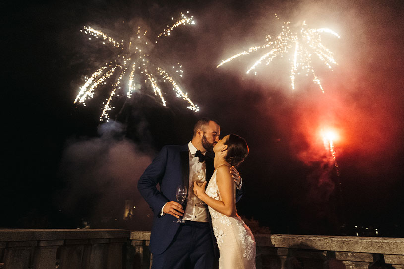 Bride and groom are kissing and holding a galss of Don Perion at Villa Bled balcony during the fireworks in night sky.