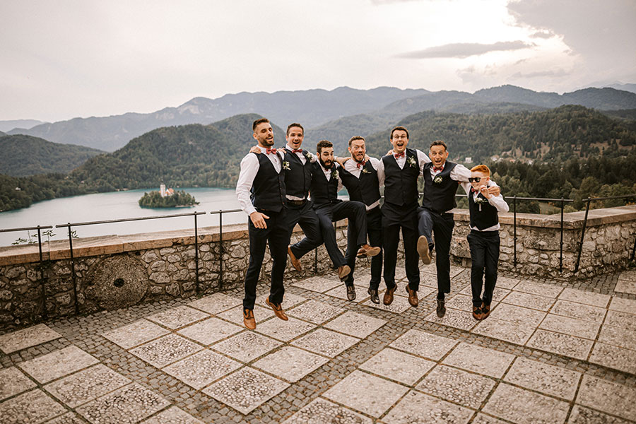 Happy groom with his best friends are jumping at Bled Castle terrace and behind them is Lake Bled Island.