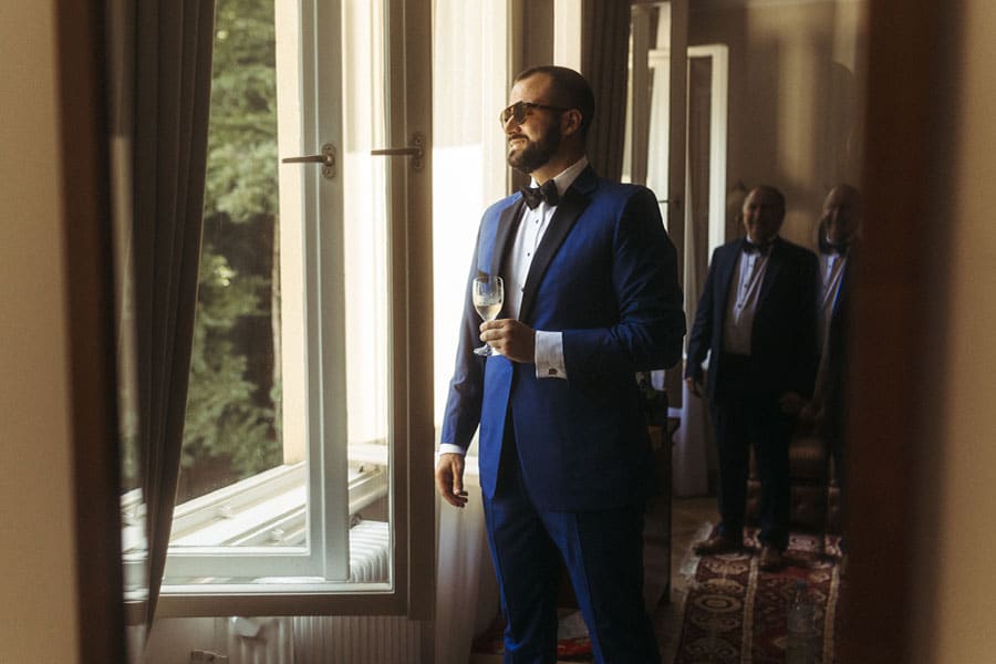 Groom in a blue suit wiht a glass of champagne is looking out the vila Bled window.