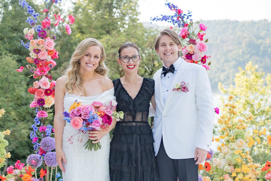 Wedding planner Petra Starbek and a happy couple under a luxurious flower arch of vibrant shades of flowers for a destination wedding in lake Bled.