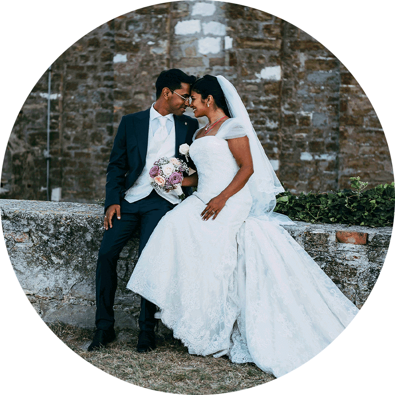 The Indian bride is in a long lace wedding dress with a pink bridal bouquet and the Indian groom are sitting on the rock fence during a wedding in SLovenia.