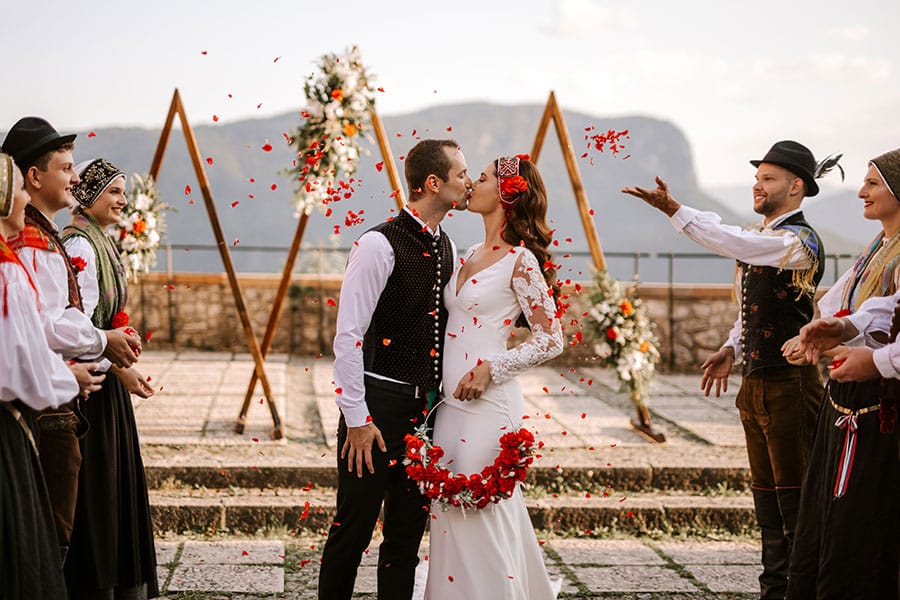 Newlyweds kisses under red fresh petals throwing by guests at their Lake Bled Castle wedding ceremony.
