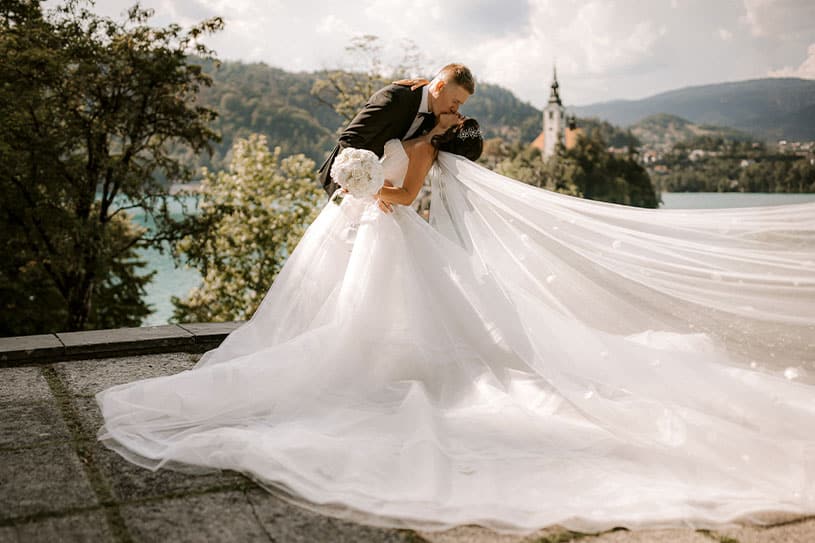 A groom and bride in a luxury wedding dress are kissing at vila Bled terrace and in back you can see magnificant island Bled with the Lake Bled Church.