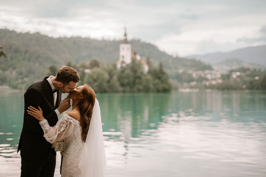 A bride in a stunning white lace wedding dress from Korea and a groom kiss on the shore of the lake Bled and behind them is Bled Island.