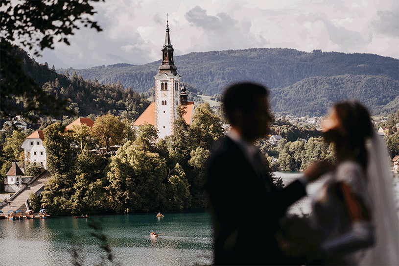 A beautiful wedding couple is standing at vila Bled bridge and behind them is the Lake Bled church.