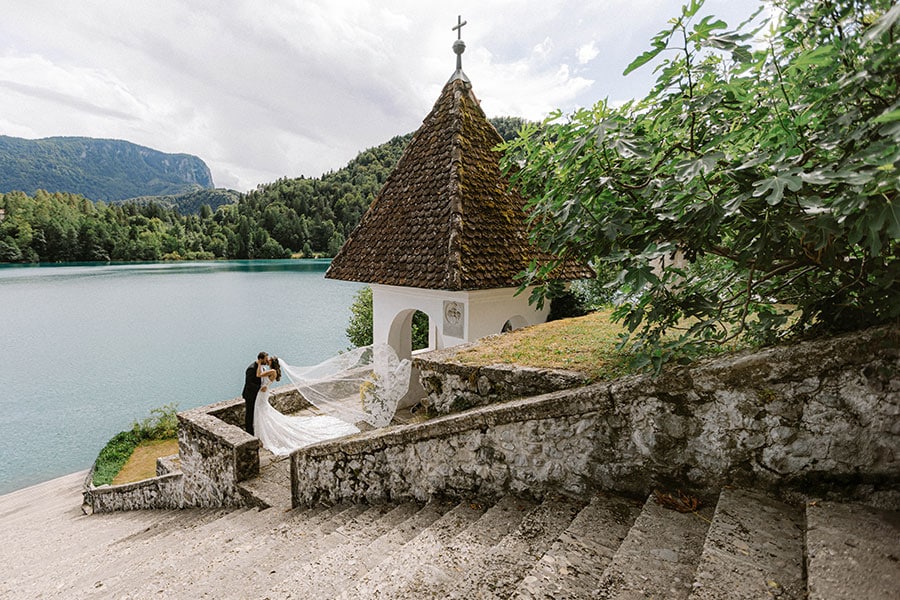 Bride and groom kissing on the historic steps of Bled Island during an intimate Lake Bled wedding 10–15 K€, with a flowing veil and lake backdrop.