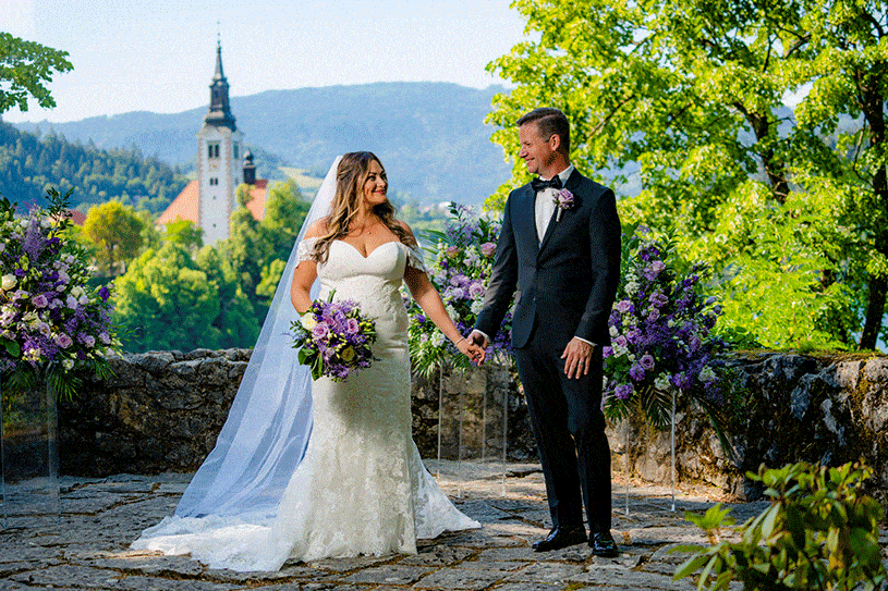 Lovely couple of newlyweds - bride and groom walking taking photos at ceremony spot decorated with purple white tall flowers at Vila Bled at Lake Bled, Slovenia.