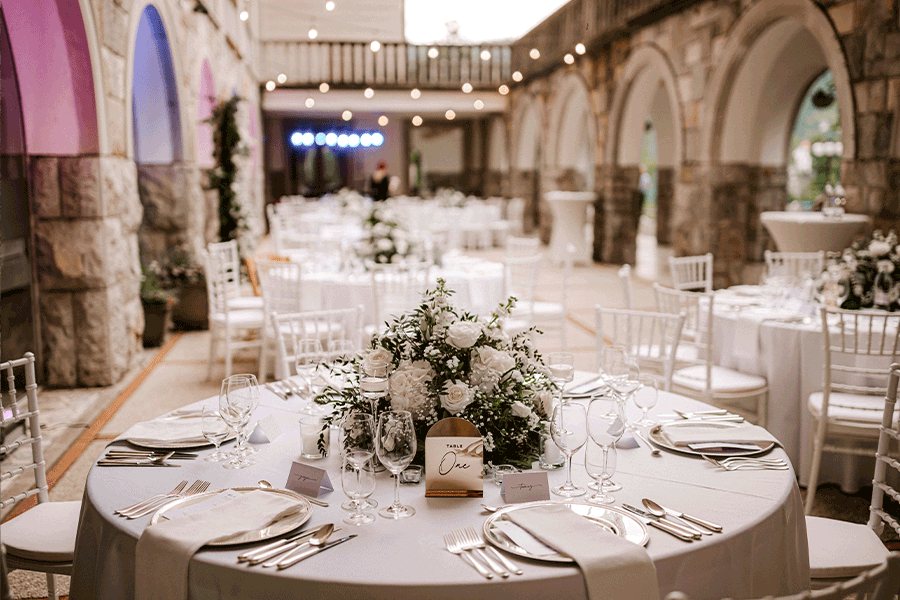 Wedding dinner set up in vila Bled with green-white flower arrangement, round tables, white wooden chairs, silver cutlery, white napkins, and gold table number.