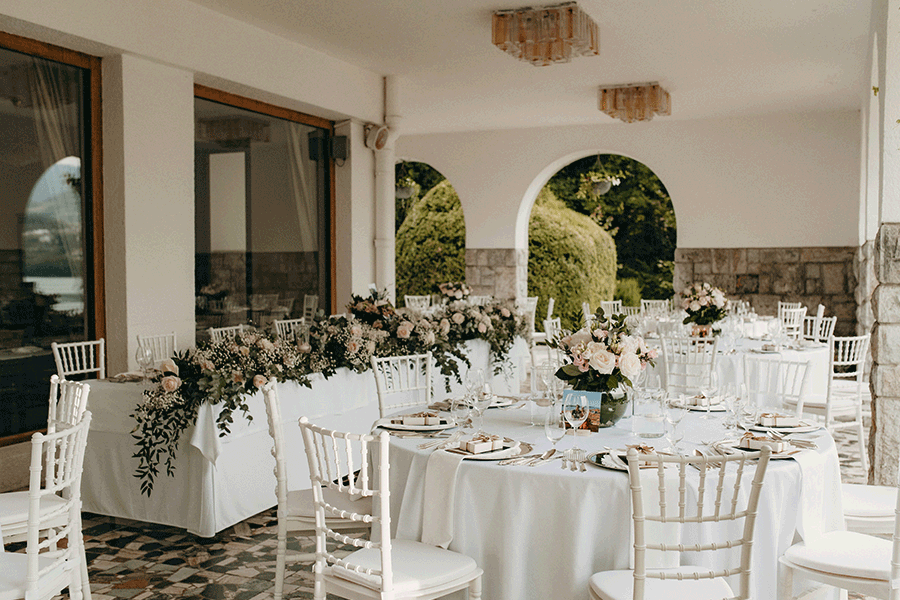 Beautiful wedding set up for a wedding dinner at vila Bled with white chairs, white table cloths, white napkins, silver underplate, silver cutlery, chocolate, pastel flower arrangements, and a picture from a couple.