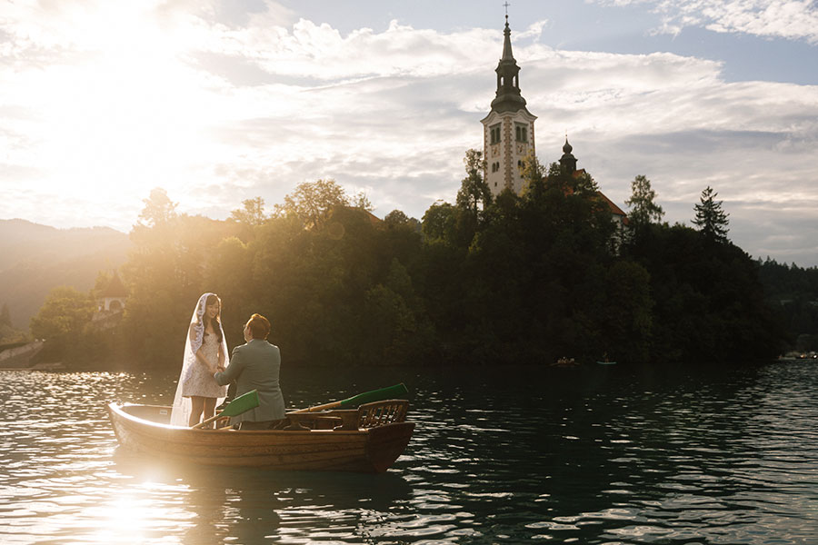 Bride and groom on a wooden boat during their Lake Bled wedding, approaching Bled Island at sunset for an intimate 10–15 K€ celebration.