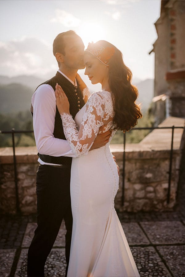 Groom give a kiss on bride's forehead at sunset az Bled Castle terrace.