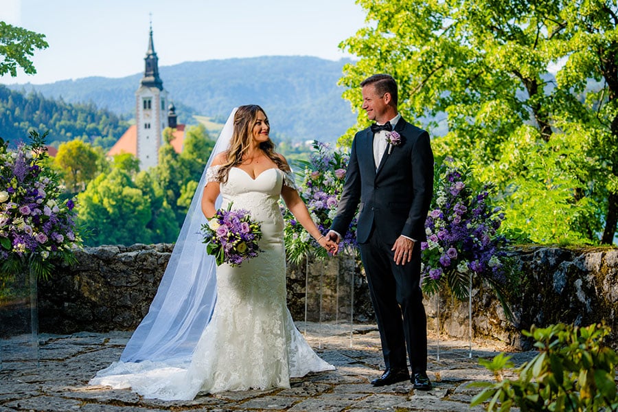 Bride in a long lace white dress with a purple wedding bouquet and groom in a black suit hold hands and stand at ceremony setting with violet flowers at vila Bled terrace.