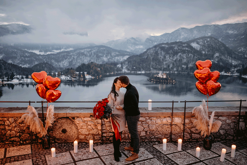 The guy is kissing his girlfriend with a red roses bouquet after she said yes in a beautiful romantic setting with red balloons and candles at Lake Bled Castle.