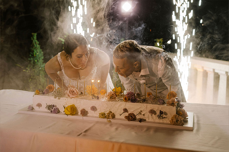 Bride and groom blowing out candles on a long rectangular wedding cake decorated with colourful flowers.