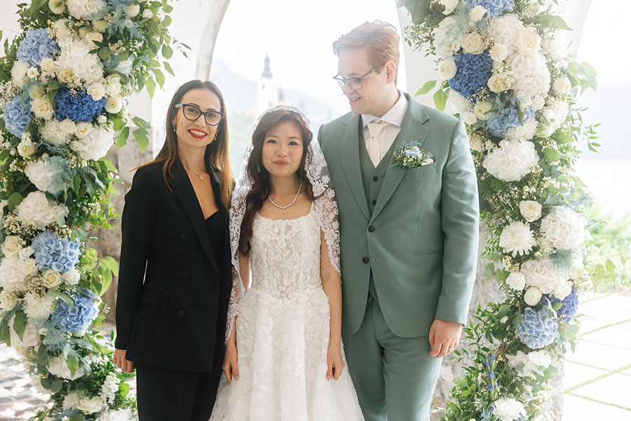 Wedding planner Petra Starbek with a couple under a floral arch at Lake Bled, guiding their bespoke wedding within a 10–15 K€ budget.