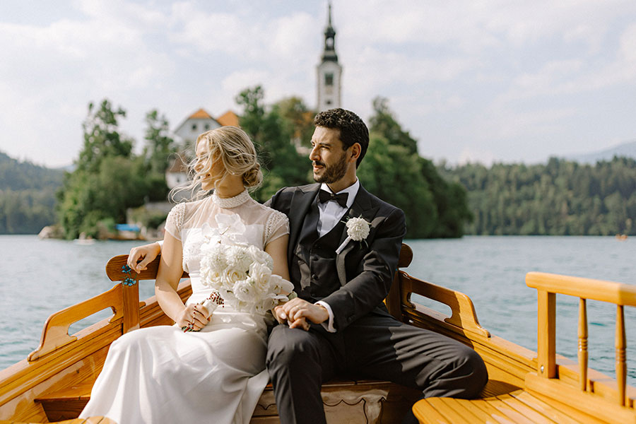 Young modern couple on a traditional wooden boat with Bled Island in the background.