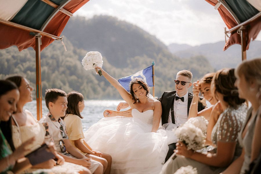 Happy guests and smiling groom with his bride who raised her white bridal bouquet during their ride on plat boat to Lake Bled Island.