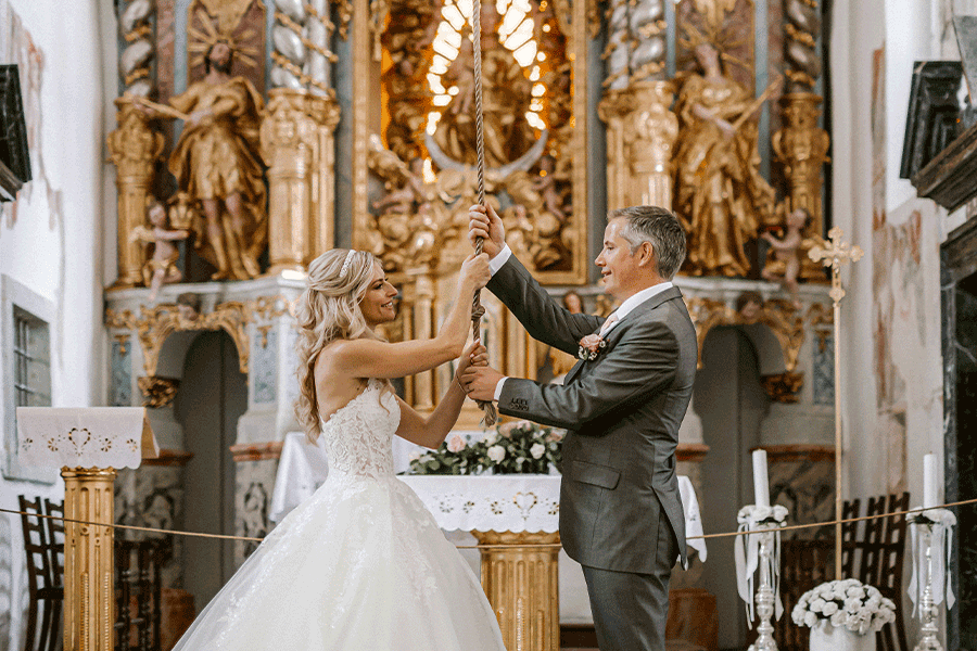 The bride in a white princess dress and the groom in a gray suit are ringing the wishing bell in the church on the Lake Bled island.