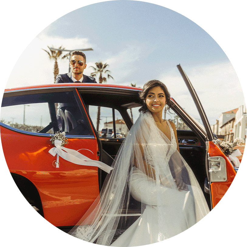 A beautiful bride in a white lace wedding dress with a royal veil and a groom in a blue suit with sunglasses are and groom with a sunglasses are going to the wedding dinner with an old red car.