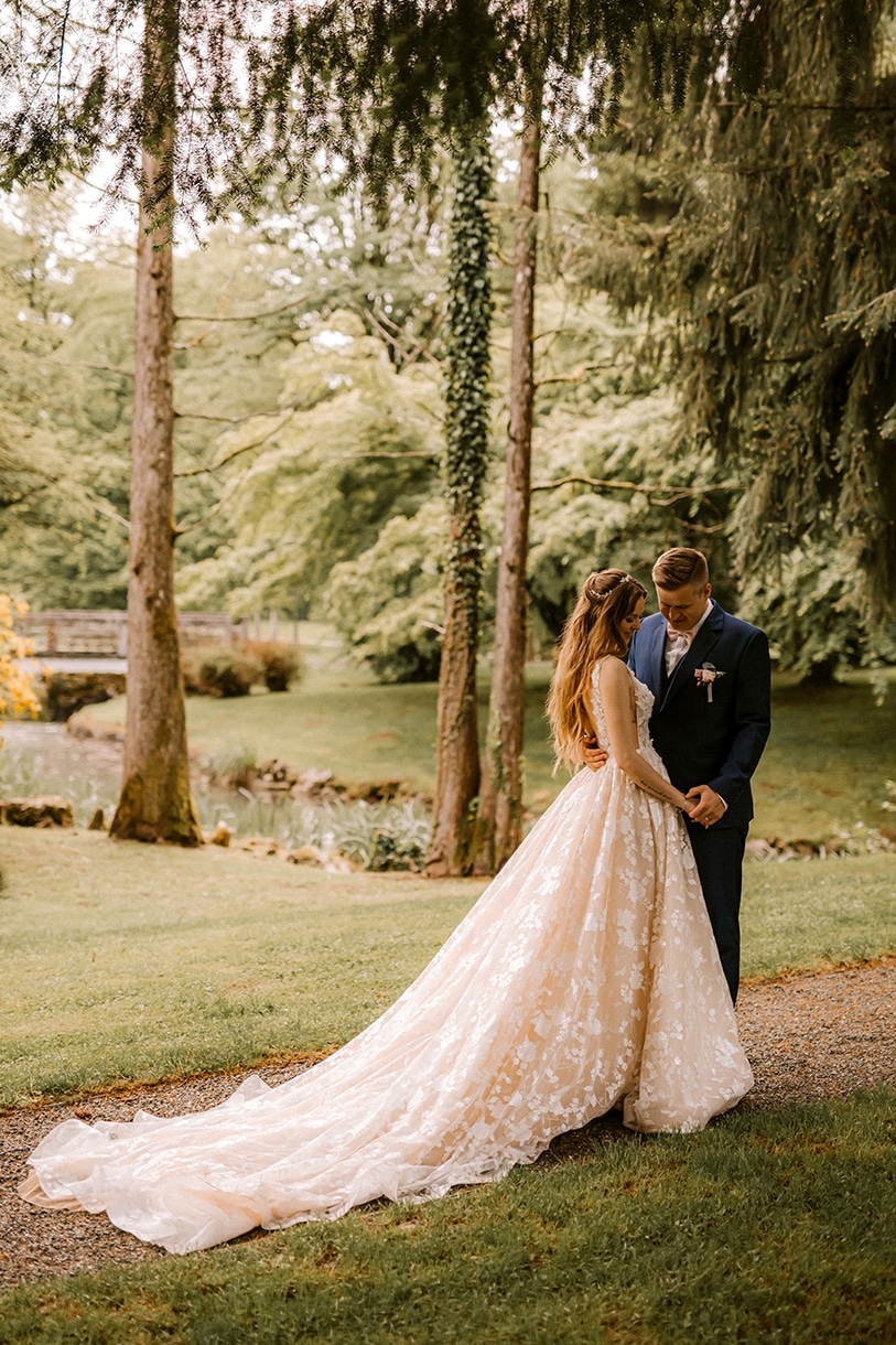 A stylish groom in a blue suit and a beautiful blonde bride in a sand lace wedding dress are standing and hugging in nature in the vila Bled park.