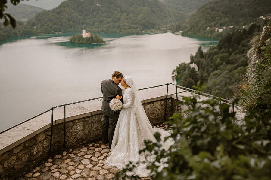A couple in love stands in their wedding dresses on the Lake Bled Castle terrace and behind them is lake Bled island.