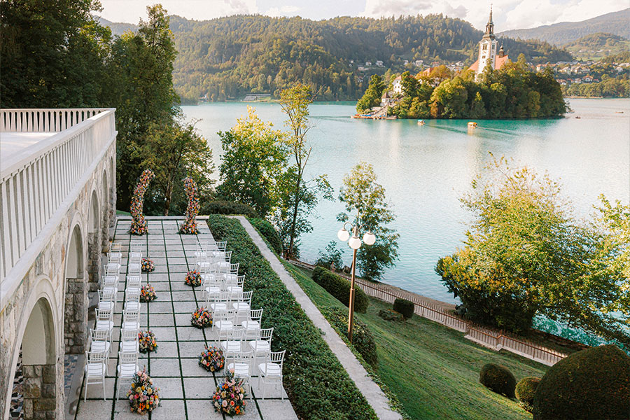 Outdoor wedding ceremony setup at Vila Bled with white chairs, colourful floral arrangements and views of Lake Bled and its island church.
