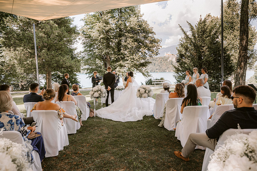 Wedding ceremony in vila Bled garden in front of the vila. White decoration, white big tall white and green flowers arrangements, bridesmaids, guests, bride in a snowwhite princess dress with royal train and groom in s black suit.