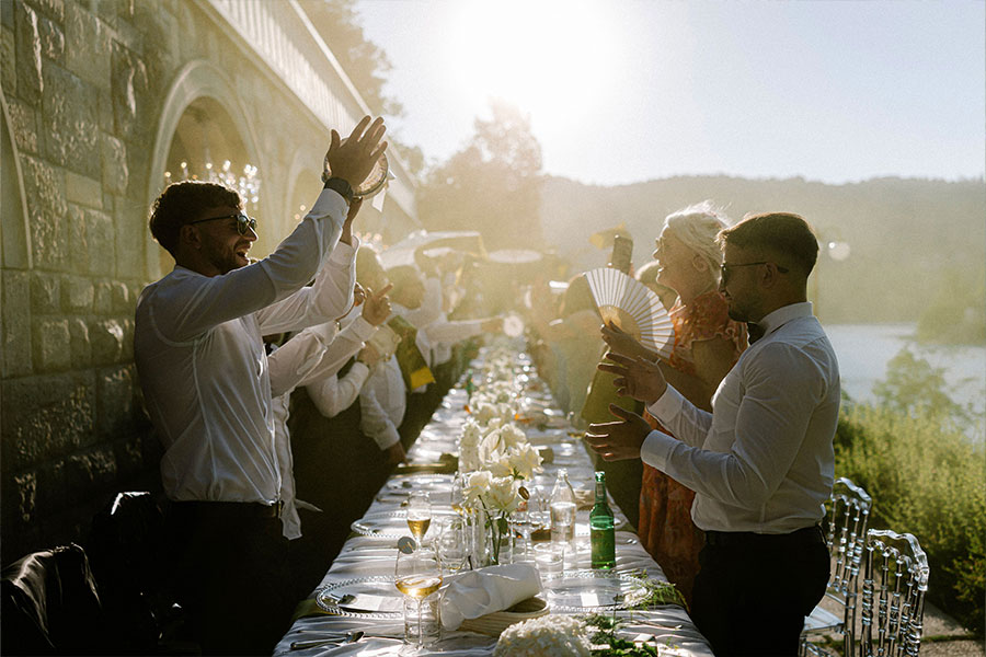 Wedding guests clapping and celebrating beside a long dining table on the Vila Bled terrace overlooking the lake.
