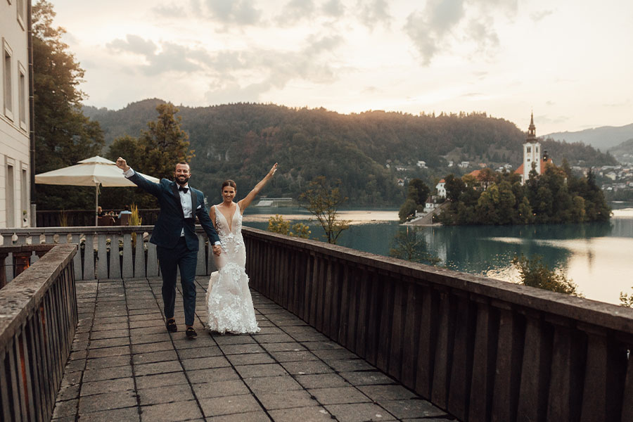 Bride in an amazing champagne lace dress and a groom in blue suit holding hands with one hand holding in the air on vila Bled terrace and behind them in a Lake bled Island.