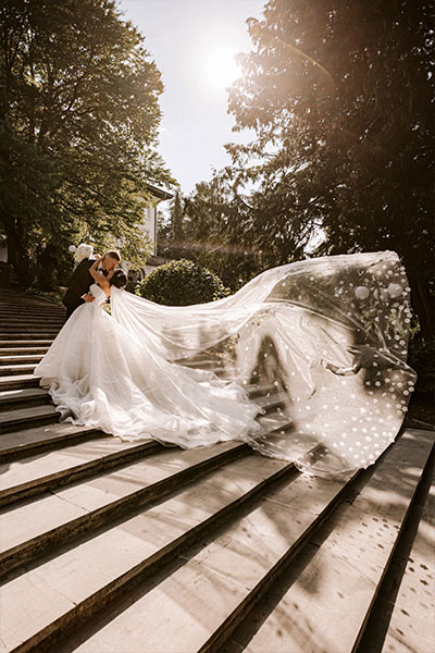 Bride in a long princess white snow wedding dress with a long veil, long trail with white bridal bouquet at vila Bled stairs and groom are kissing at Vila Bled stairs.