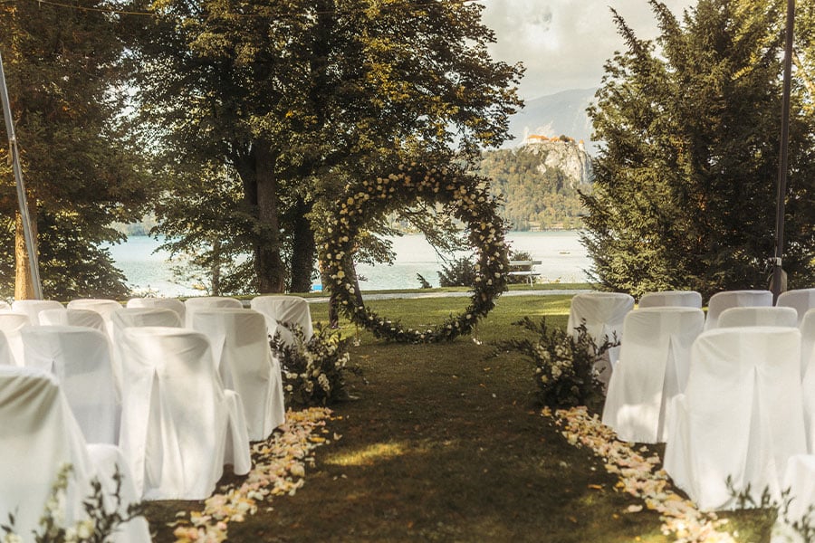 Wedding ceremony setting in Vila Bled park with an amazing circle flower arch and white chair cover chairs. Next to the chairs are fresh white petals and flower arrangements. 