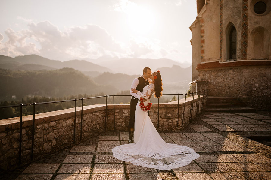 A bride in a long white wedding dress and a groom in black suit are kissing at lake Bled Castle terrace with a beautiful view of lake Bled and mountains in sunset.